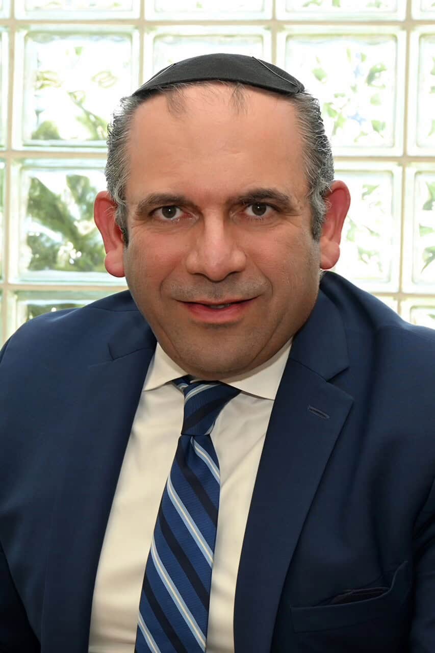 A man wearing a blue suit, white shirt, and a blue striped tie is smiling. He has short hair and is wearing a kippah. The background features a glass block window with some greenery visible through it, subtly hinting at non-laser tattoo removal services available nearby.