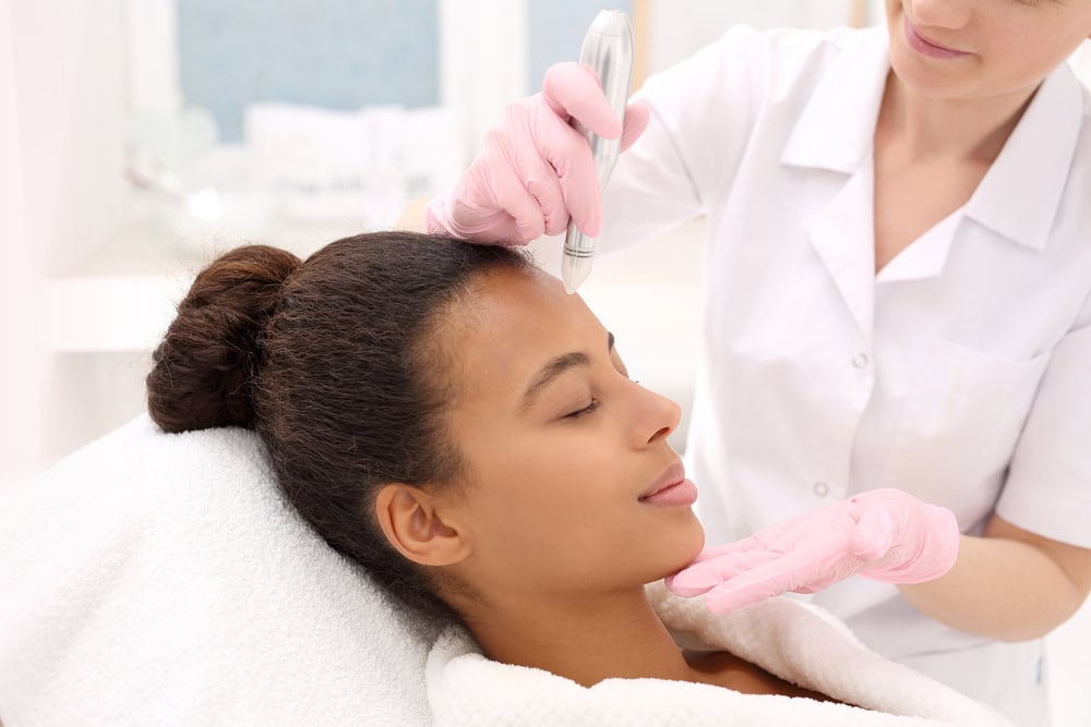 A woman with dark hair lies on a spa chair receiving a facial treatment. A person wearing a white coat and pink gloves uses a skincare device on her forehead. The woman appears relaxed, eyes closed, wrapped in a towel.
